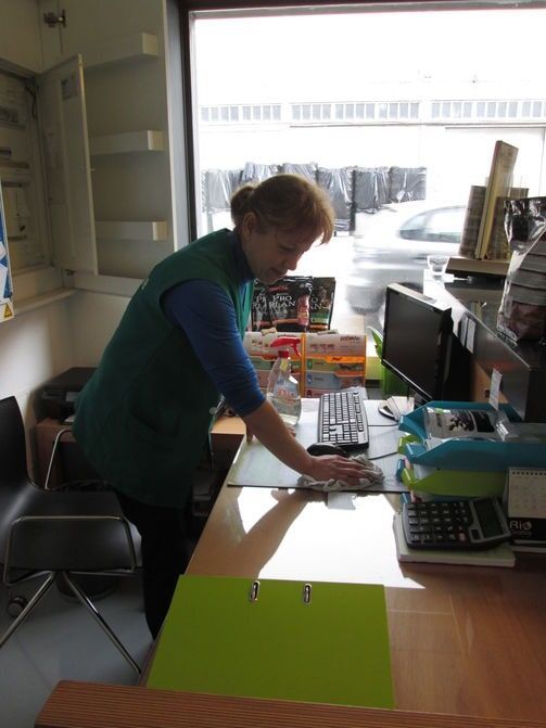Mulher limpando um balcão em uma loja; produtos, computador e calculadora sobre a mesa.