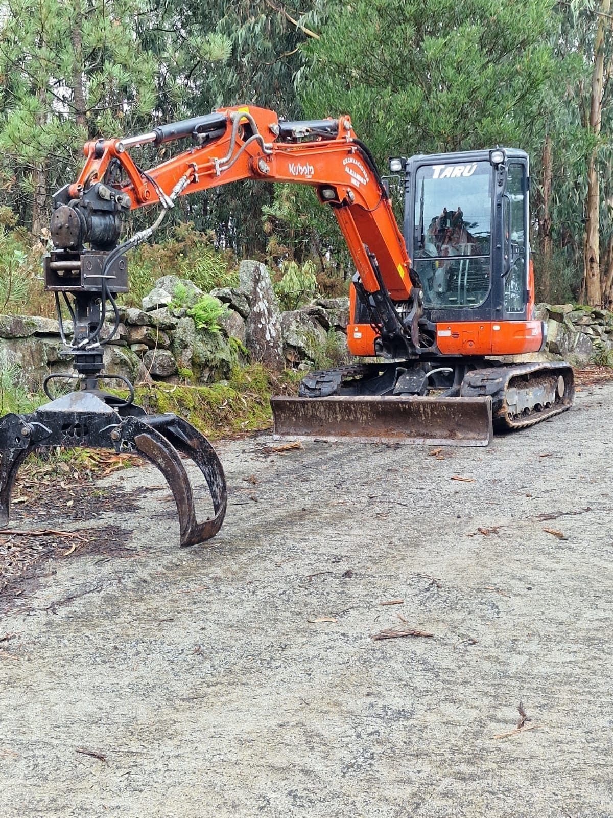 Excavadora naranja con garra estacionada en un camino de grava junto a un terraplén rocoso.