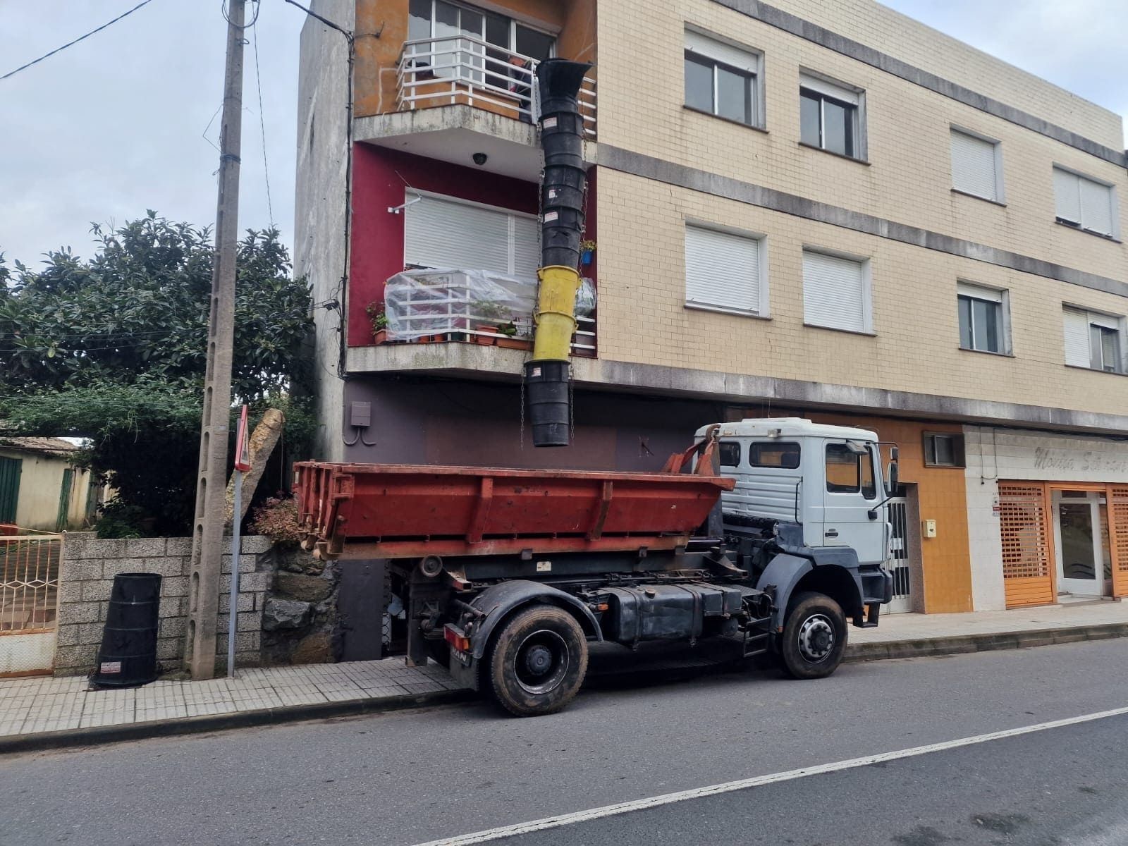 Un camión volquete rojo estacionado junto a un edificio de apartamentos beige con balcones en una calle de la ciudad.