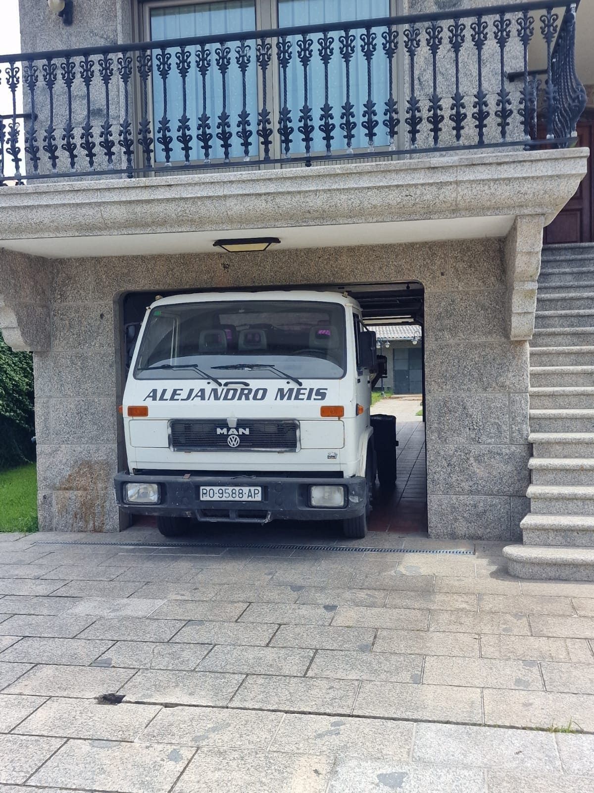 Camioneta blanca estacionada en un garaje debajo de un balcón, con un camino de entrada de piedra y una barandilla azul encima.
