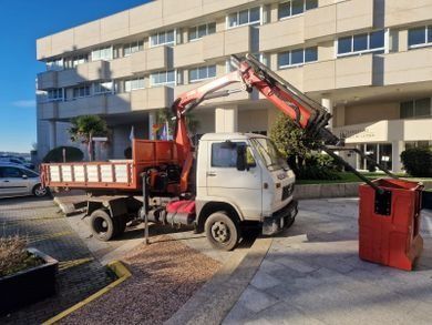 Un camión grúa rojo estacionado junto a un edificio beige con un contenedor de basura rojo enfrente.
