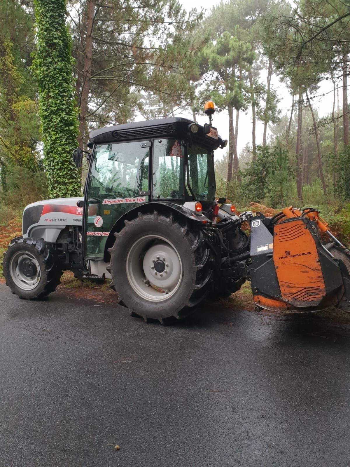 Tractor gris con quitanieves naranja en un camino mojado en una zona boscosa durante la lluvia.