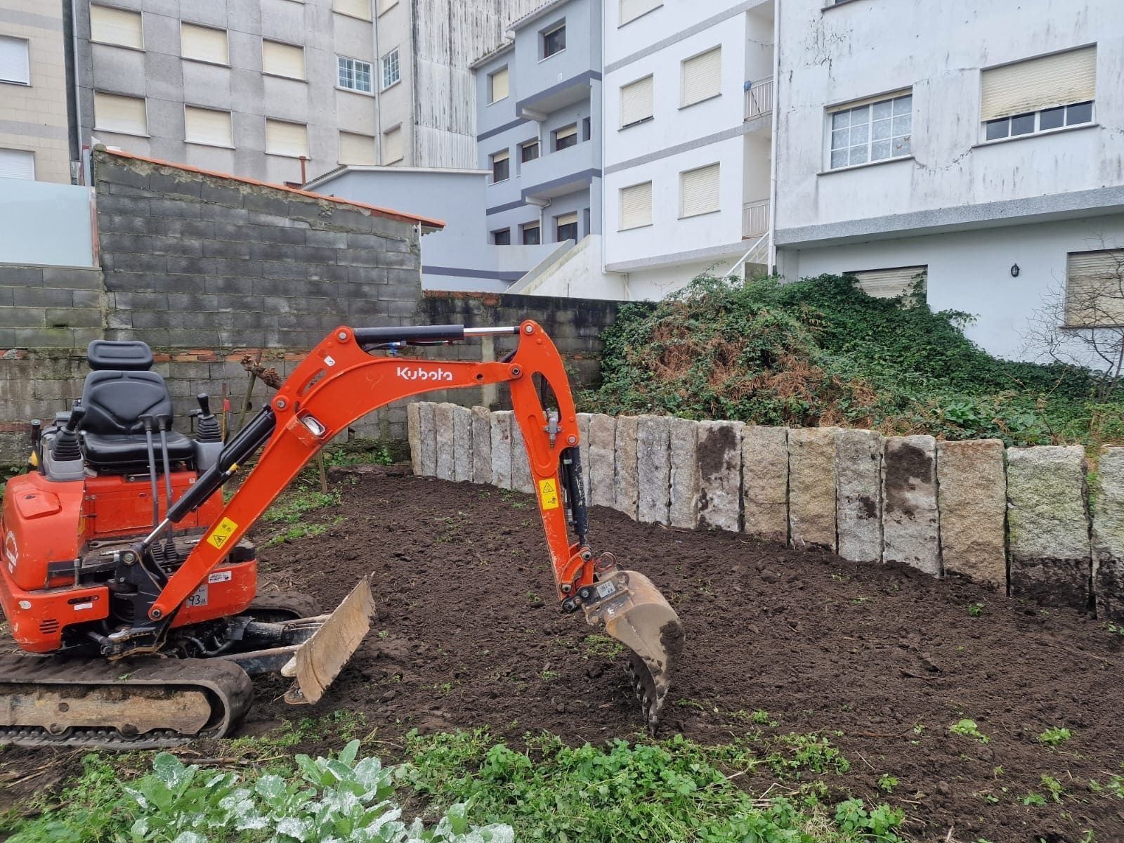 Una excavadora naranja está removiendo tierra oscura junto a edificios de apartamentos y un muro de piedra.