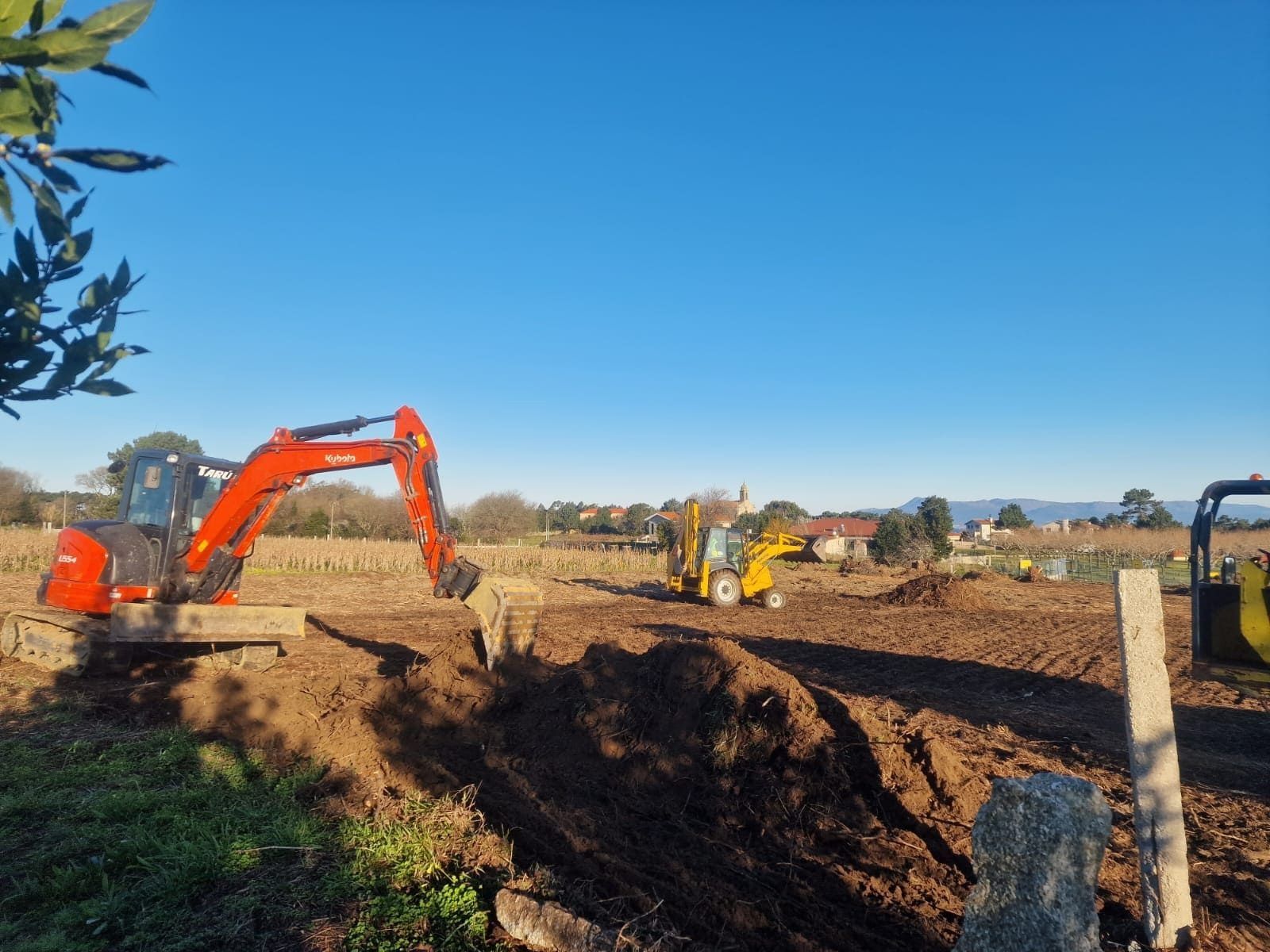 Una excavadora naranja excava en una obra fangosa junto a una máquina amarilla bajo un cielo azul.