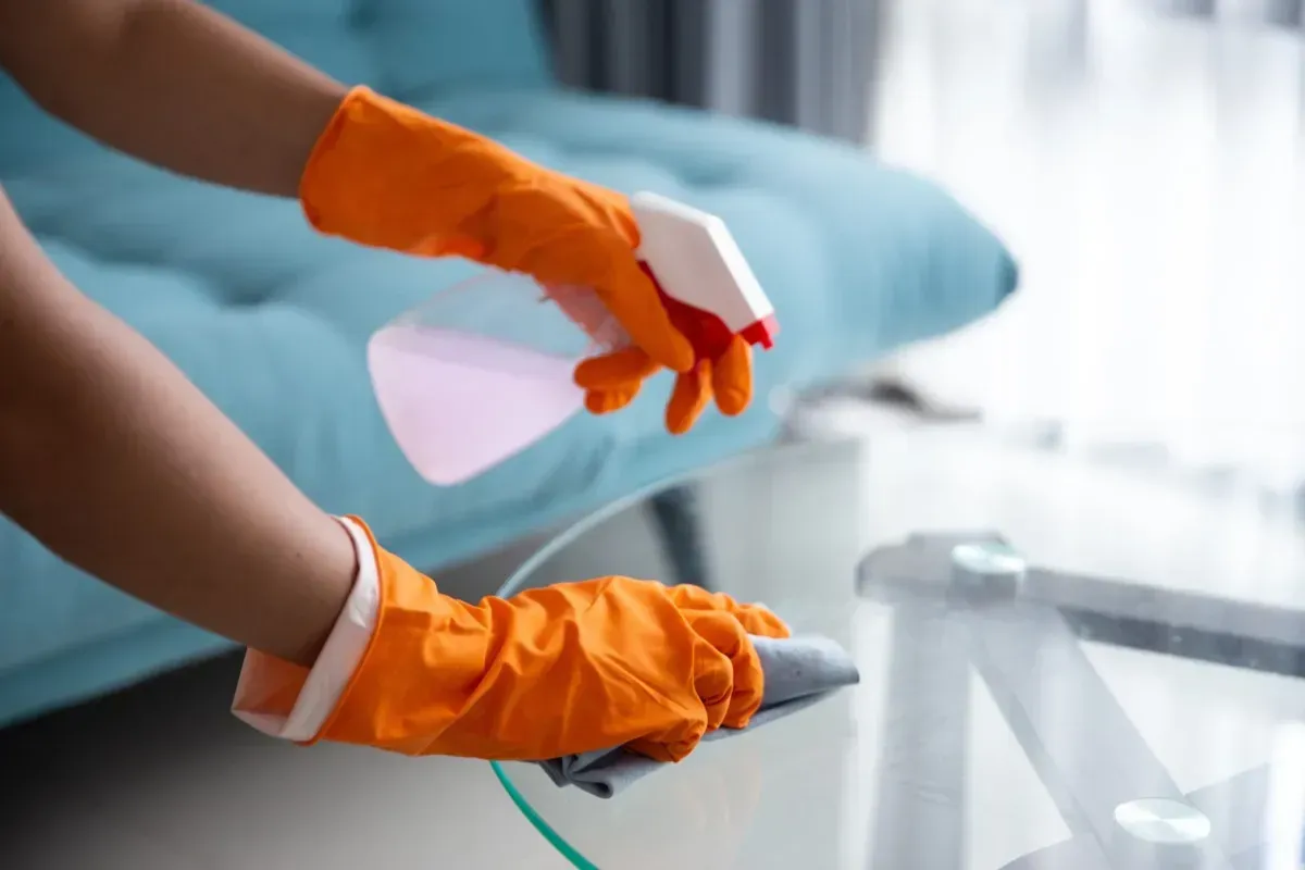 Persona con guantes naranjas, rociando y limpiando una mesa de vidrio con un paño.