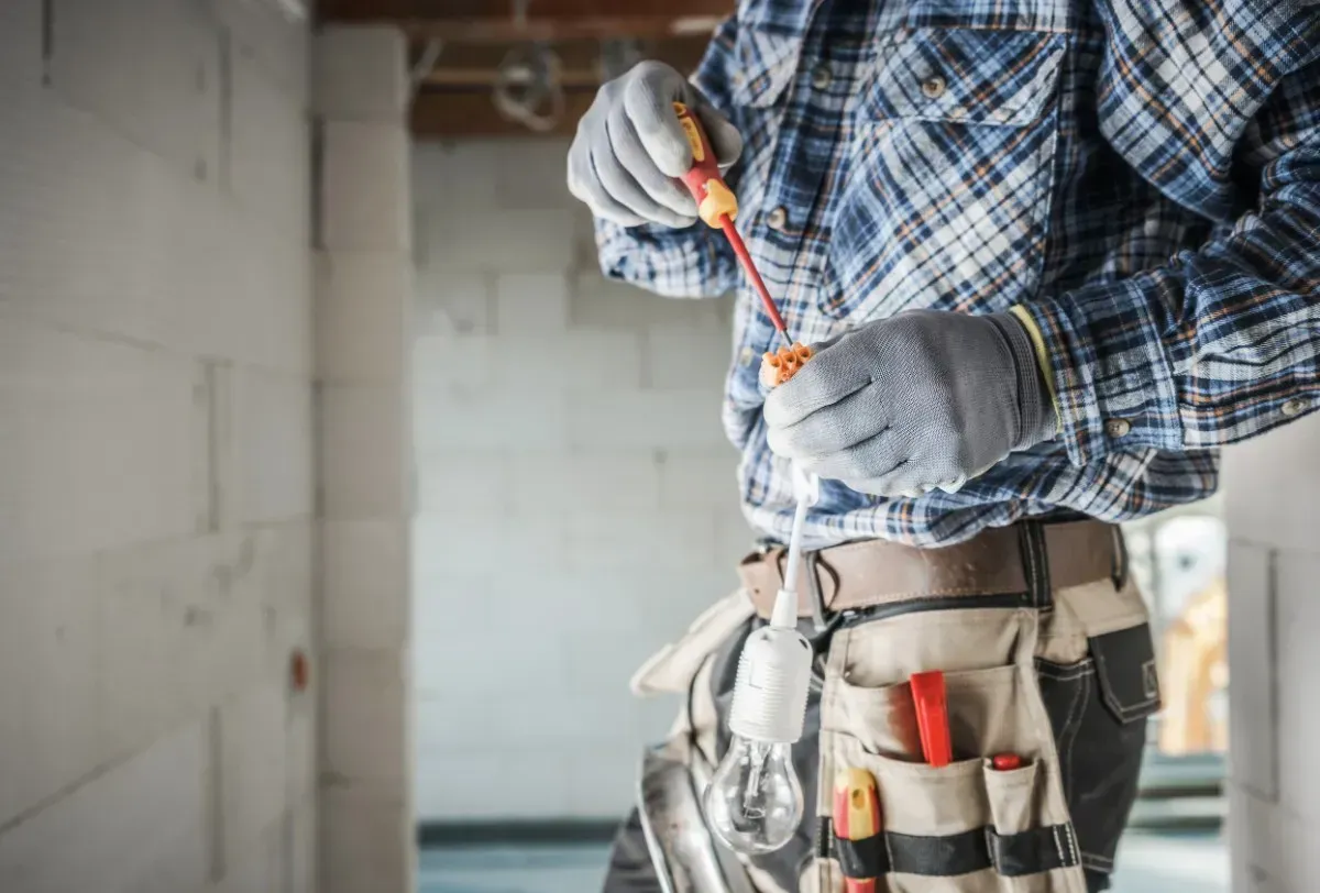Electricista trabajando, sosteniendo un destornillador, usando cinturón de herramientas y guantes en un área de construcción.