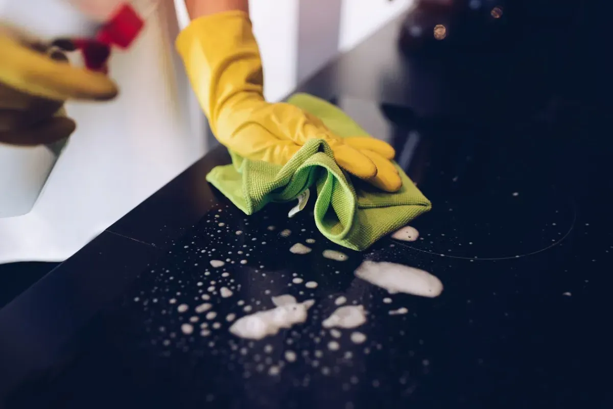 Una persona que lleva guantes amarillos rocía y limpia una superficie oscura con un paño verde.
