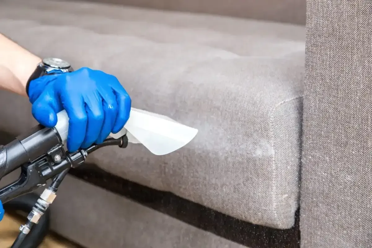 Persona con guantes azules limpiando un sofá gris con una herramienta de limpieza.