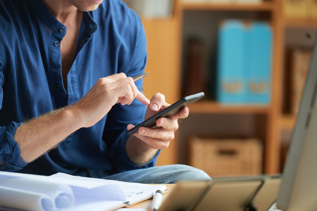 Persona con camisa azul usando un teléfono inteligente en un escritorio con papeles y una computadora portátil cerca.