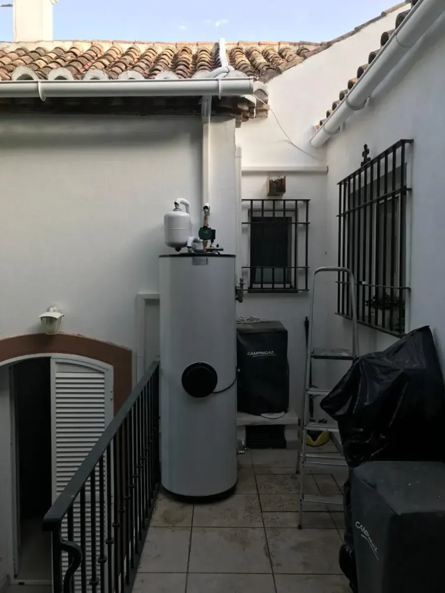 Un alto tanque blanco de agua caliente se alza sobre un patio embaldosado entre dos muros blancos de un edificio