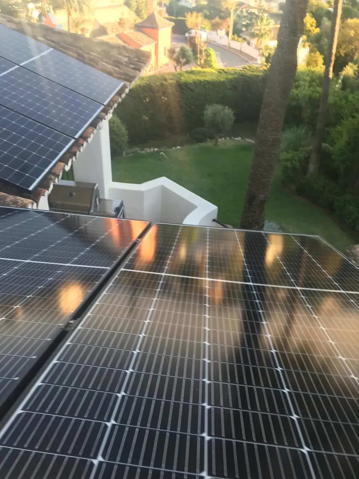 Paneles solares instalados en un tejado inclinado con vistas a un patio trasero con césped, árboles y un barrio residencial.