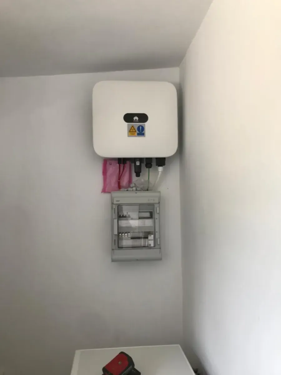 Un inversor solar Huawei blanco montado en la pared, encima de una pequeña caja de fusibles gris, en una habitación.