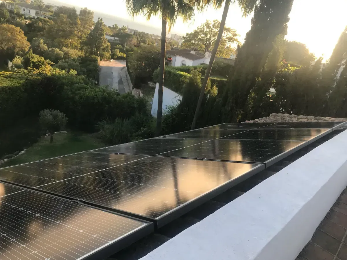 Una hilera de paneles solares negros instalados en un tejado blanco, con vistas a un jardín y árboles iluminados