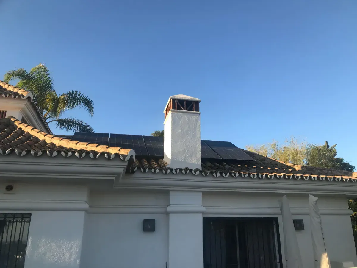 Una casa de estuco blanco con techo de tejas, paneles solares y una chimenea sin terminar, contra un cielo azul despejado.