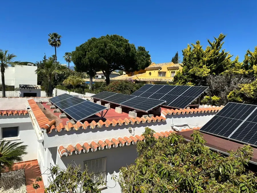 Hileras de paneles solares instalados en el tejado de una casa con borde de tejas rojas, rodeada de árboles