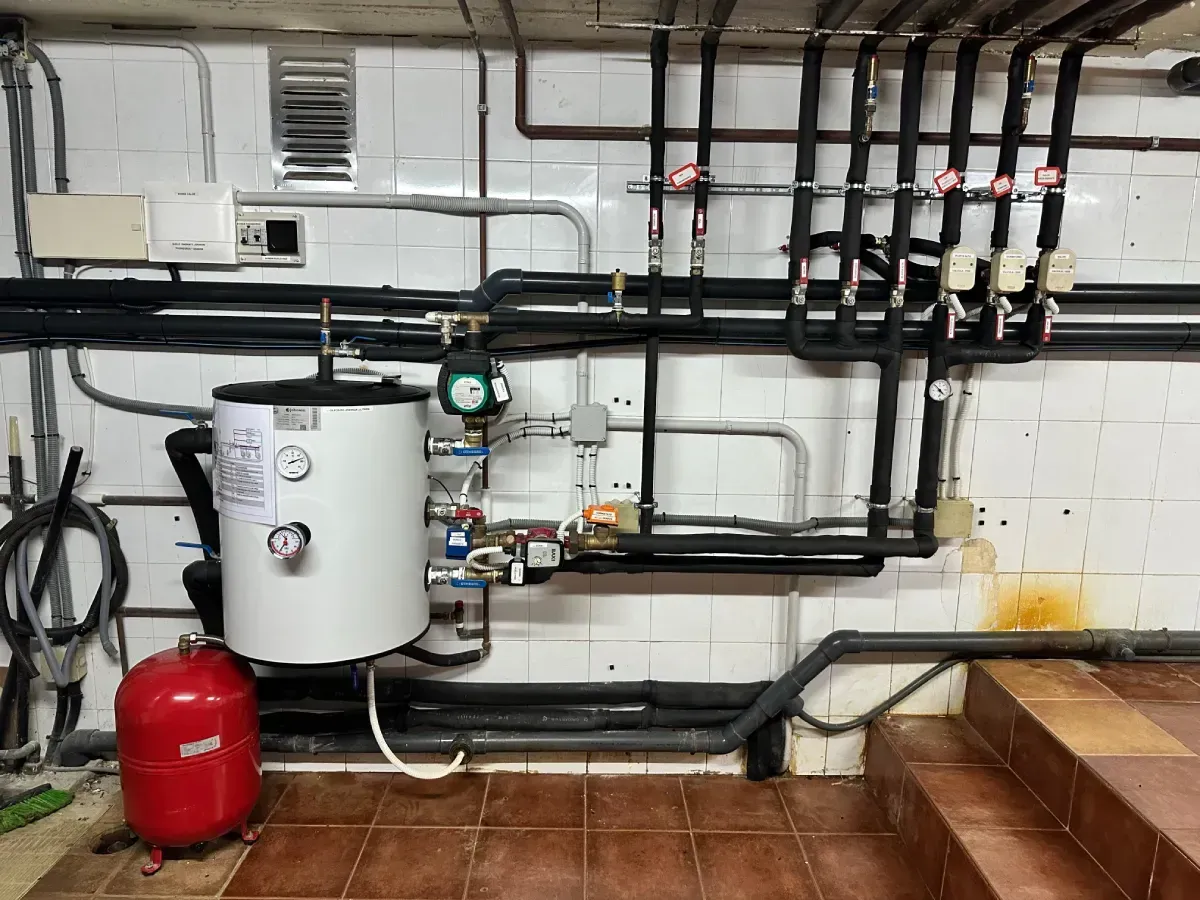 Un depósito de agua caliente blanco y un vaso de expansión rojo montados sobre una pared alicatada con una red de tuberías