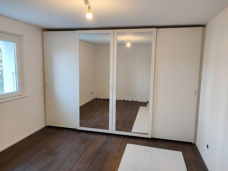 Camera da letto vuota con pareti bianche, pavimento in legno scuro, ante dell'armadio a specchio e una finestra sulla sinistra.