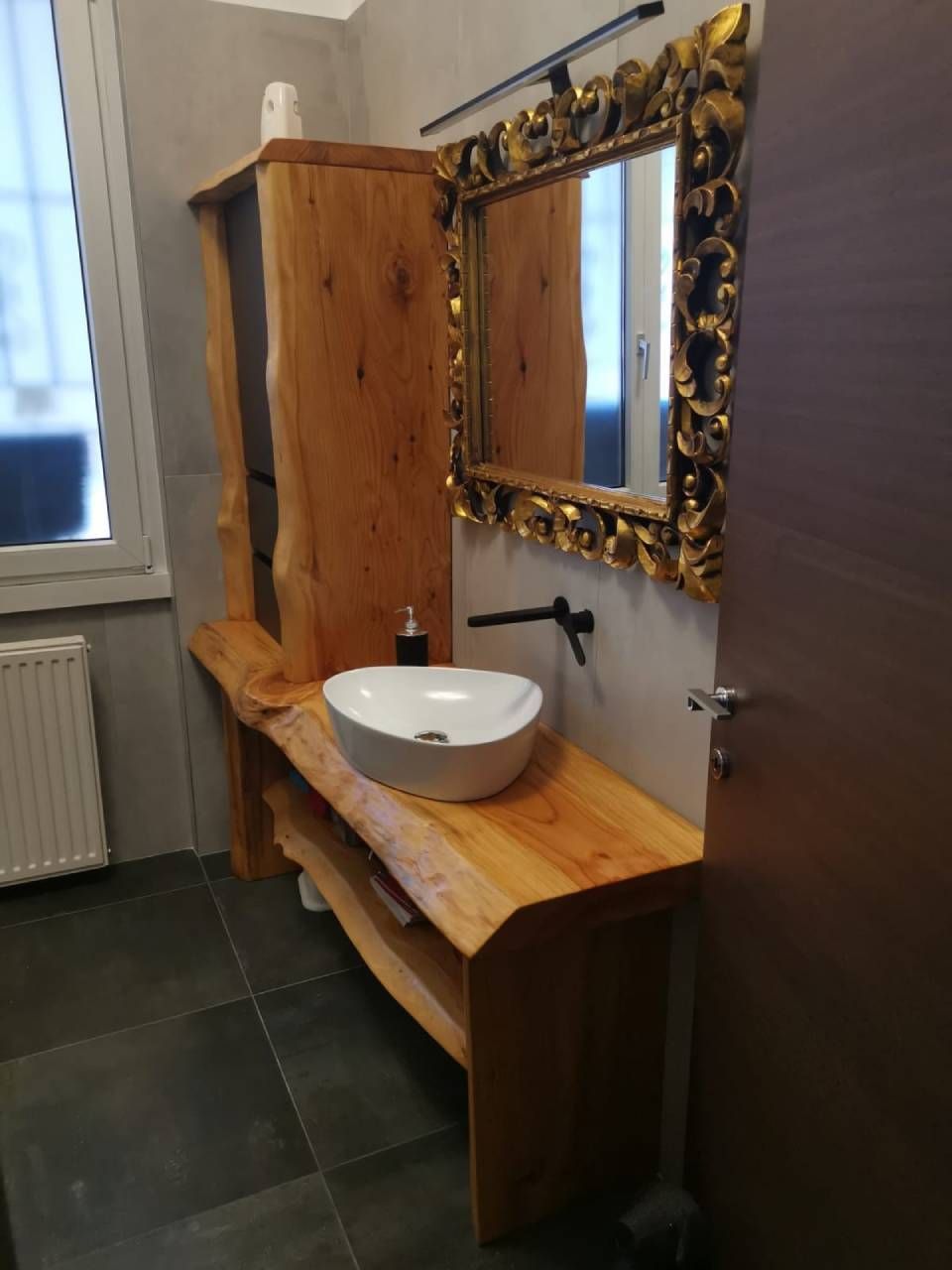 Mobile da bagno in legno con lavabo rotondo bianco e specchio decorato con cornice dorata, posizionato contro una parete piastrellata.