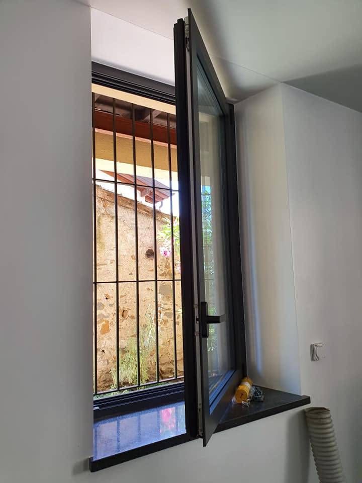 Finestra aperta con sbarre nere, che mostra un muro di pietra e un tetto all'esterno, accanto a un muro grigio e a un tubo di sfiato.