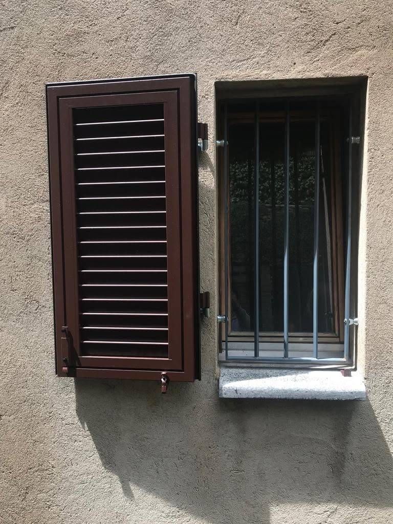 Parete esterna con due finestre adiacenti, una con persiane marroni e l'altra con inferriate metalliche.