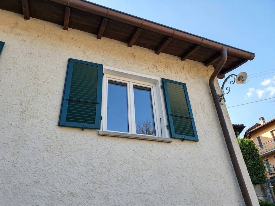 Esterno della casa con muro in stucco bianco, persiane blu e una finestra centrale sotto una grondaia marrone.