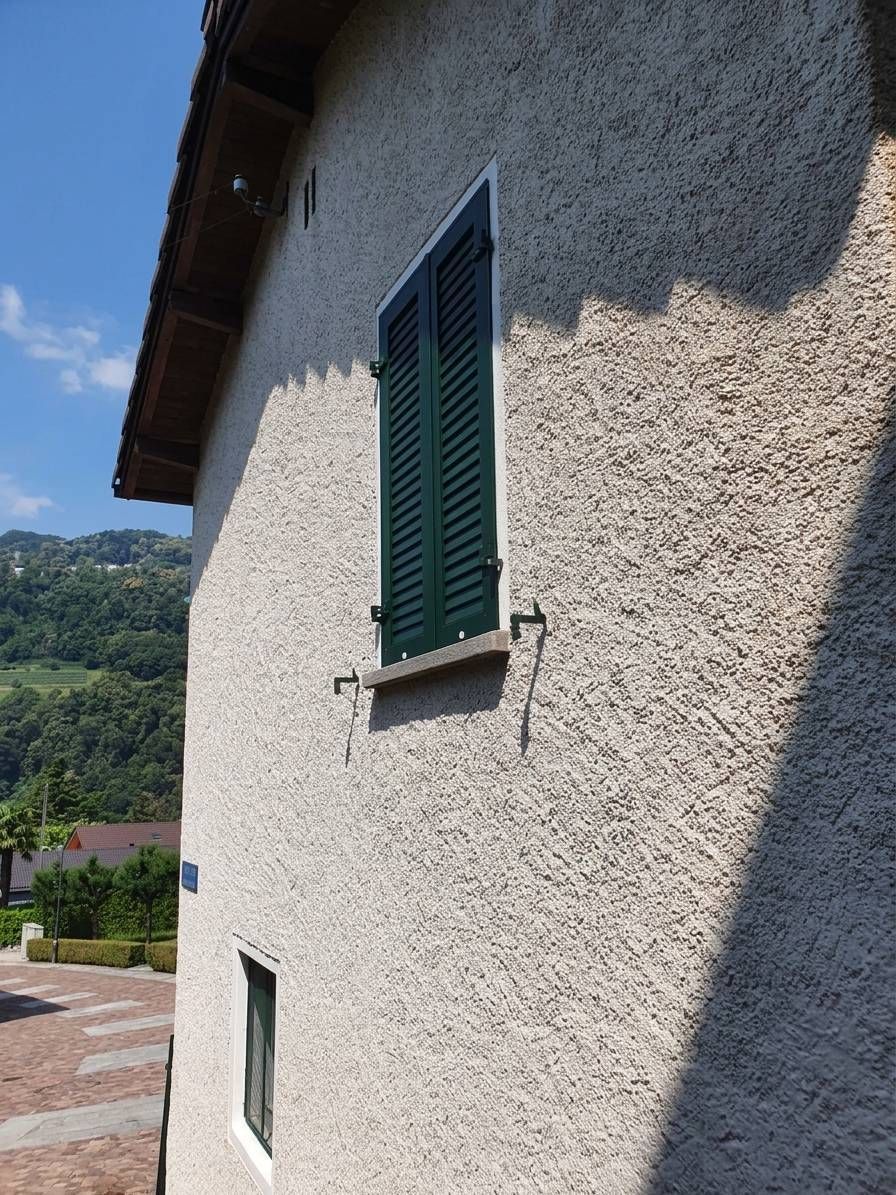 Muro di una casa intonacato di bianco con una finestra con persiane verdi, illuminata dal sole, accanto a una strada di montagna.