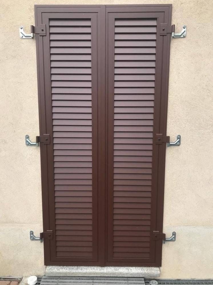 Persiane a lamelle marroni chiuse su una parete in stucco beige, fissate con staffe metalliche.