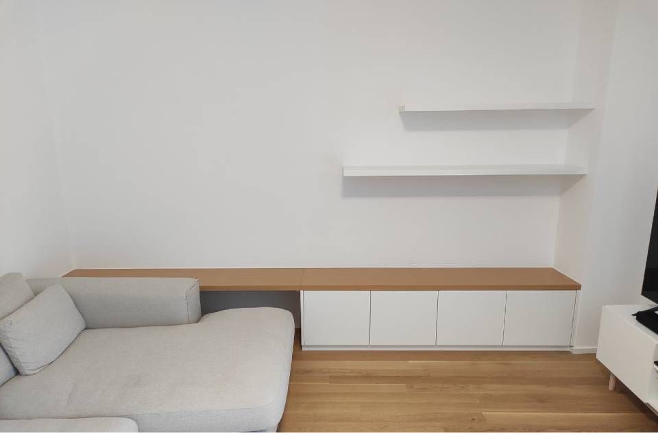 Soggiorno minimalista con divano grigio chiaro, pavimento in legno e mensole a muro bianche.