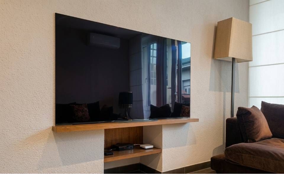 Soggiorno moderno con TV a schermo piatto montata a parete, mensola in legno chiaro, lampada da terra e divano marrone.