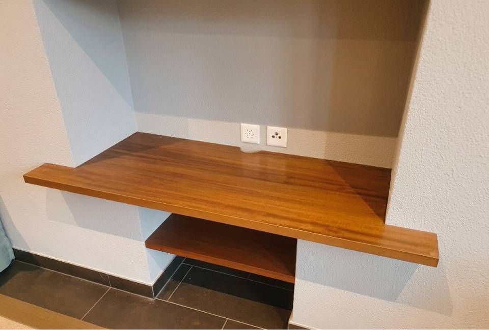 Angolo scrivania in legno integrato con prese a muro e ripiano inferiore in una stanza piastrellata.