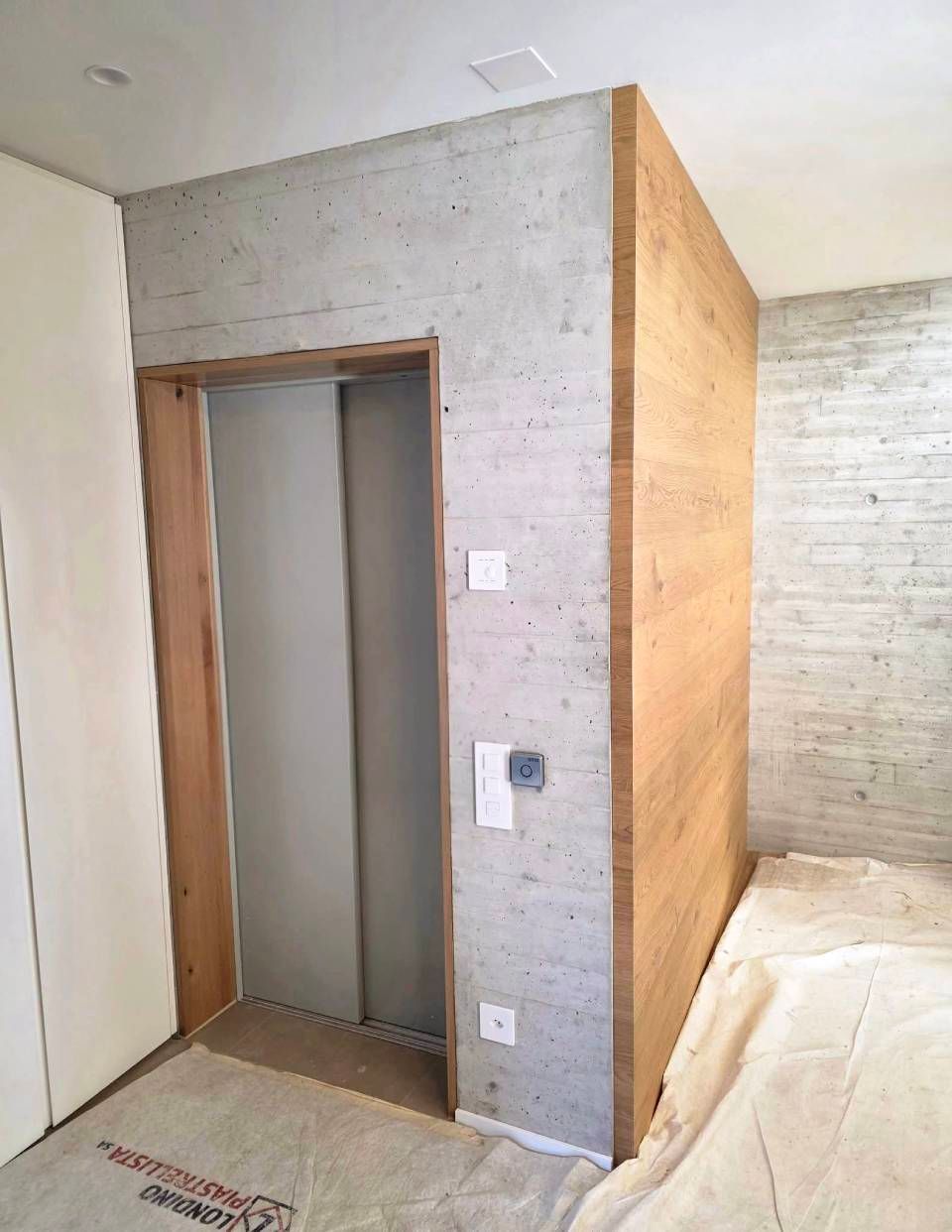Interno non rifinito con apertura metallica per l'ascensore, pareti in cemento e pannelli in legno.