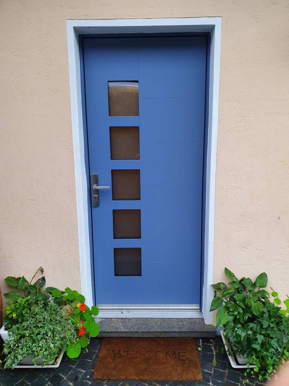 Porta d'ingresso blu con quattro finestre quadrate, incorniciate da piante in vaso e uno zerbino.