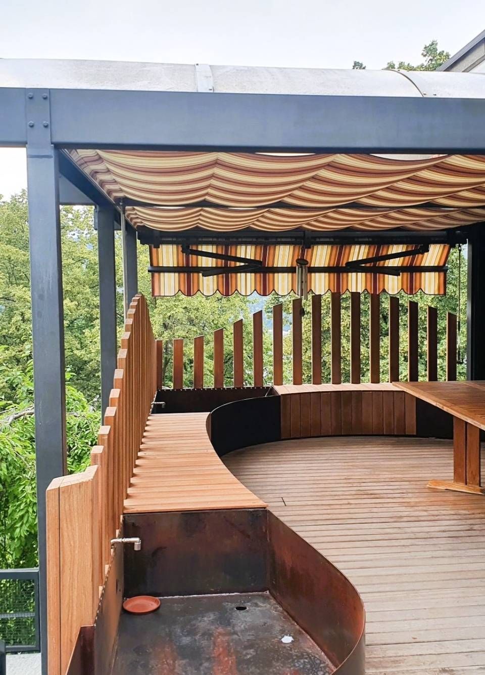 Area esterna coperta in legno con panca curva e tavolo sotto una tettoia a righe.