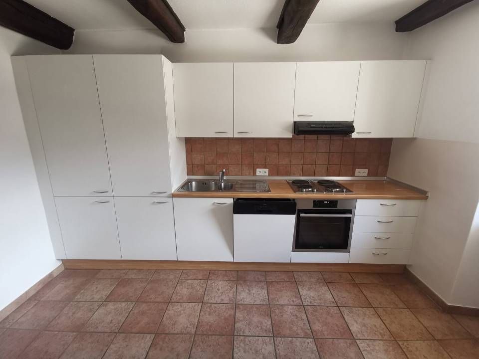 Piccola cucina bianca con travi in legno, pavimento piastrellato e mobili moderni, lavello, piano cottura e forno.