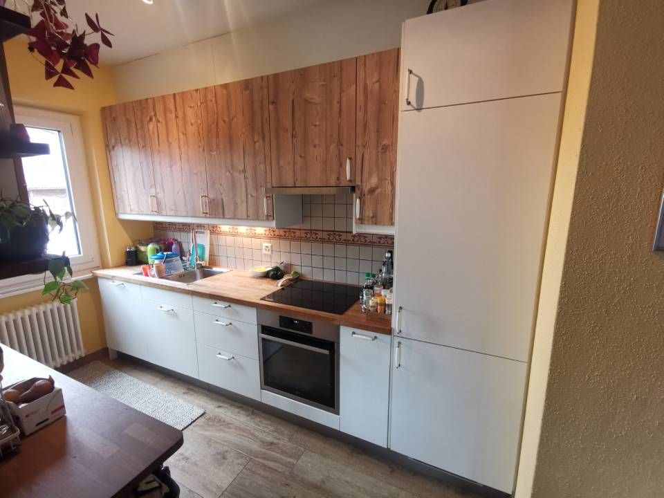 Piccola cucina con mobili in legno, pensili bassi bianchi, forno e un frigorifero bianco alto accanto a una finestra soleggiata.