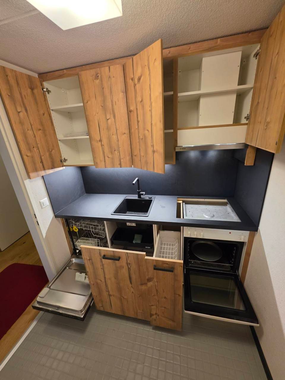 Cucina piccola con mobili in legno, piano di lavoro nero, lavello, fornelli ed elettrodomestici compatti.