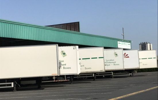 Des camions blancs stationnés sous l'auvent d'un bâtiment vert par une journée ensoleillée.