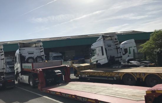 Plusieurs semi-remorques blanches sur des remorques à plateau, stationnées près d'un bâtiment par une journée ensoleillée.
