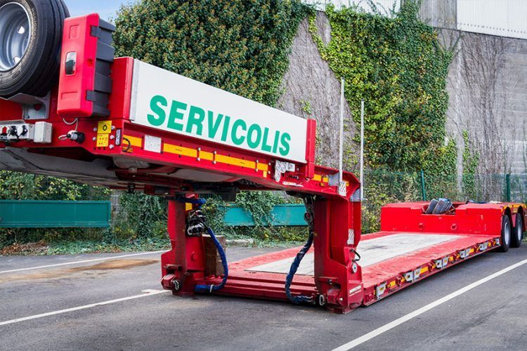 Remorque plateau rouge Servicolis sur une route goudronnée avec de la végétation verte en arrière-plan.