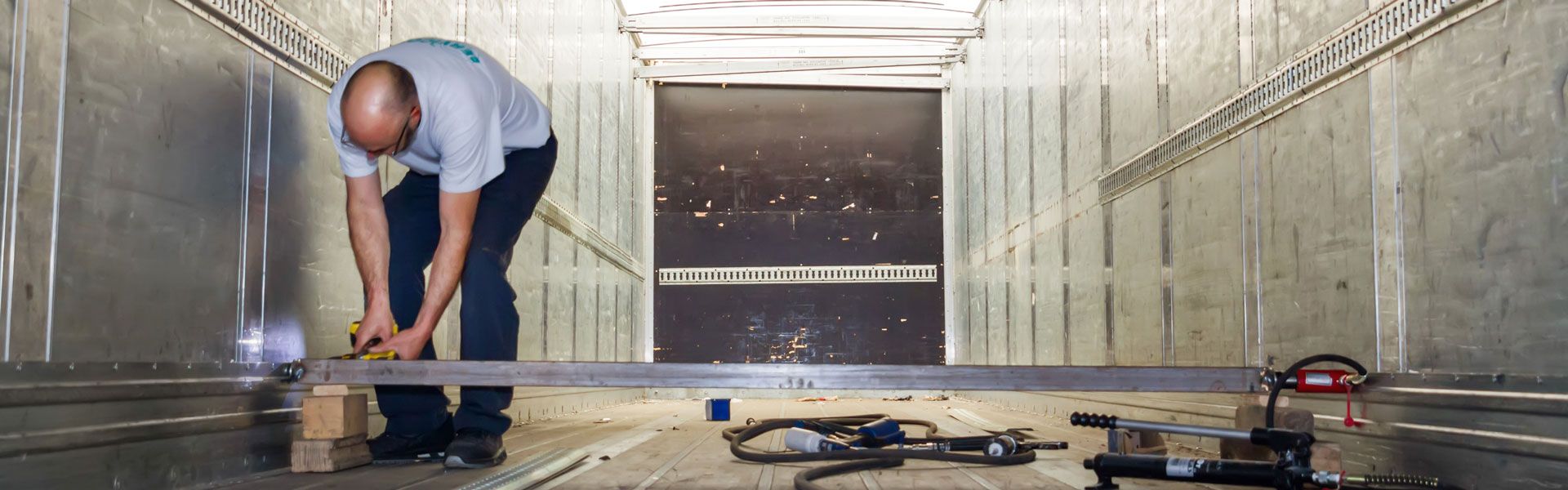 Un homme travaille à l'intérieur d'une boîte métallique. Il est penché, en train de mesurer ou de couper quelque chose près du sol.