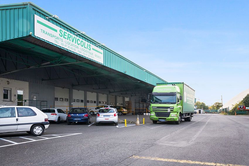Camion vert à quai de chargement, portant l'inscription « Servicolis ». Voitures garées à proximité. Ciel bleu.