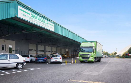 Un camion vert est stationné à un quai de chargement, le bâtiment portant l'enseigne SERVICOLIS. Des voitures sont garées à proximité.