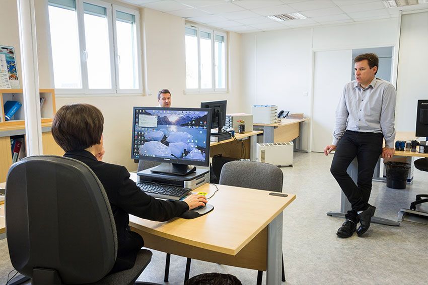 Dans un bureau, trois personnes sont présentes. L'une est assise à un bureau et utilise un ordinateur, une autre observe la scène, et la troisième est assise à un bureau en arrière-plan.