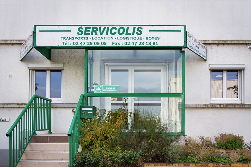 Façade du magasin SERVICOLIS avec auvent vert, escalier et paroi vitrée ; bâtiment blanc.