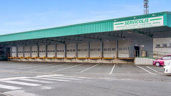 Extérieur d'un entrepôt Servicolis avec plusieurs quais de chargement. Auvent vert au-dessus des quais, voitures stationnées.
