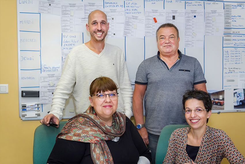Quatre personnes sourient devant un tableau blanc. Deux sont debout, deux sont assises. Elles portent des vêtements beiges, bleus et noirs.