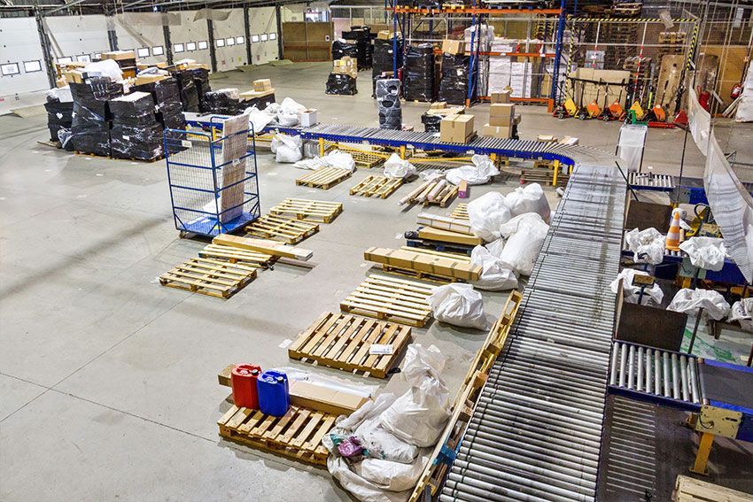 Intérieur d'entrepôt avec palettes, tapis roulants, cartons et un chariot bleu.