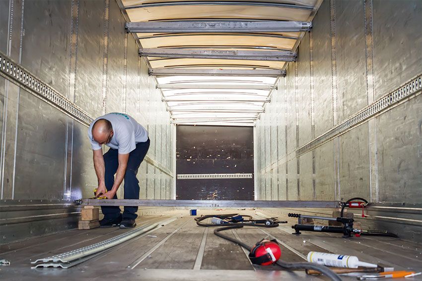 Un homme à l'intérieur d'une remorque prend des mesures à l'aide d'outils posés au sol. Vue intérieure : parois métalliques, plafond en bois.