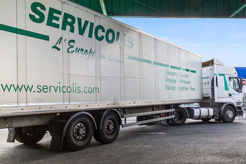 Semi-remorque Servicolis blanche avec logo vert garée sous un pont ; journée ensoleillée.