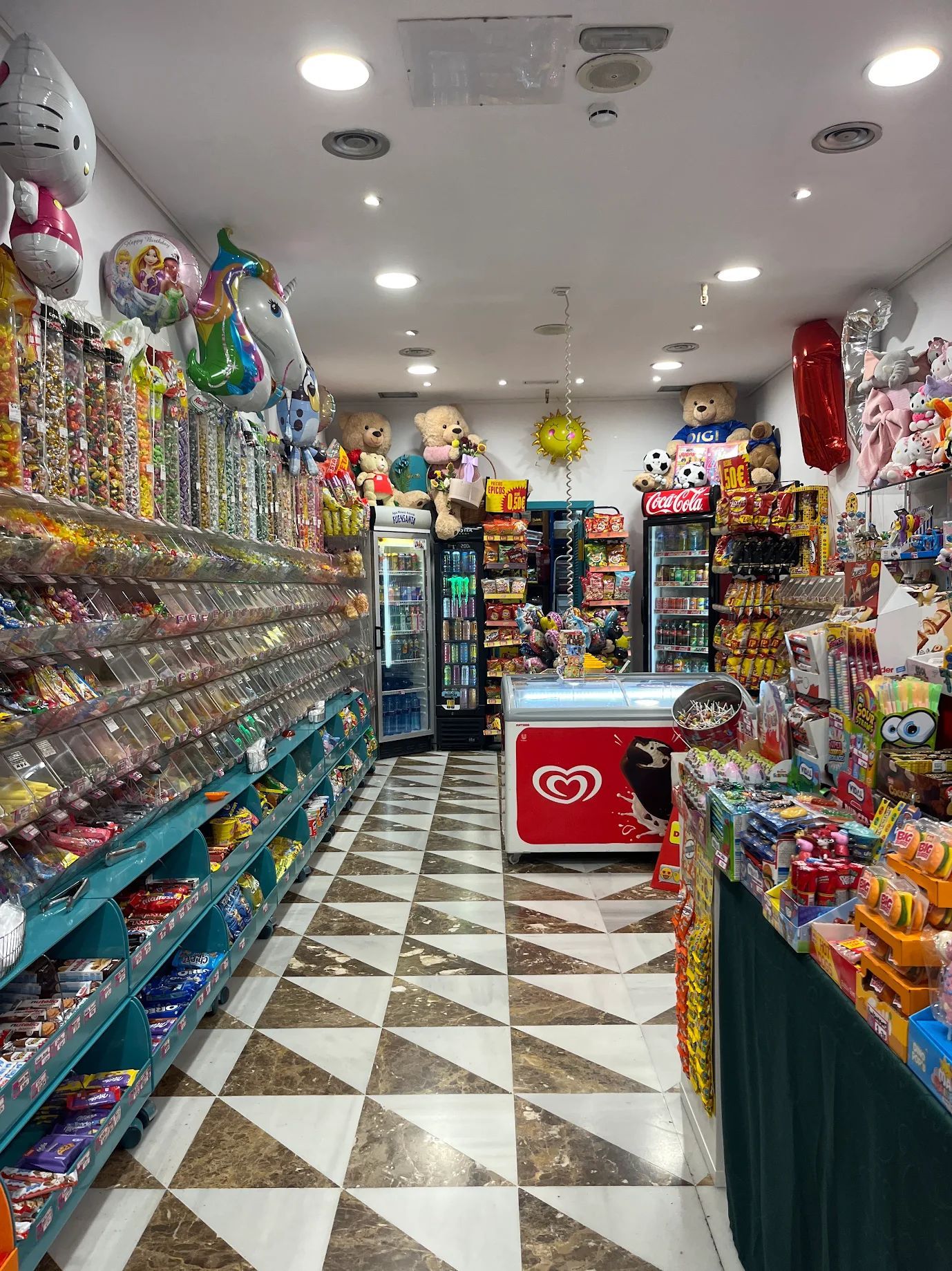 Interior de tienda de dulces lleno de estantes con golosinas de colores, globos y congelador de helados.